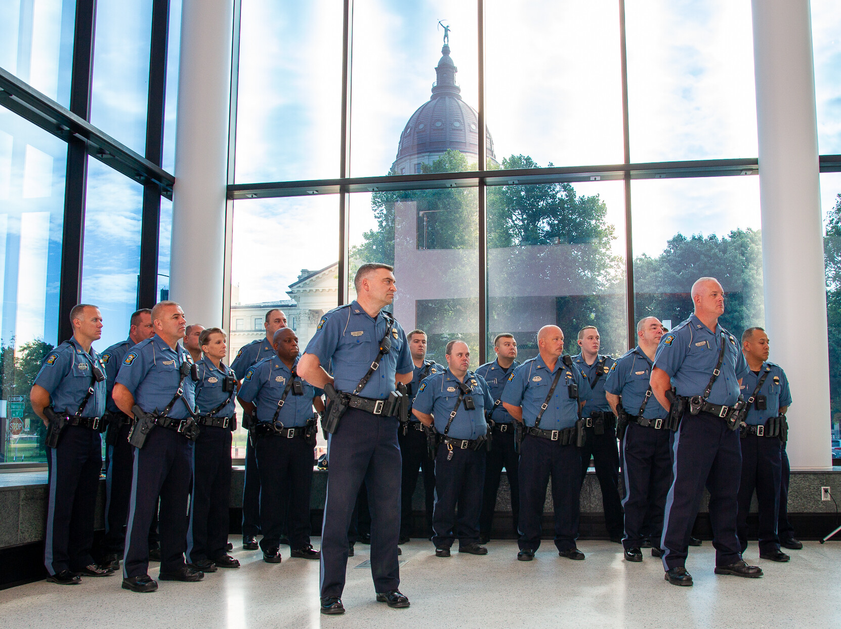 Group of troopers stand in front of a bank of windows with the capitol building in the background